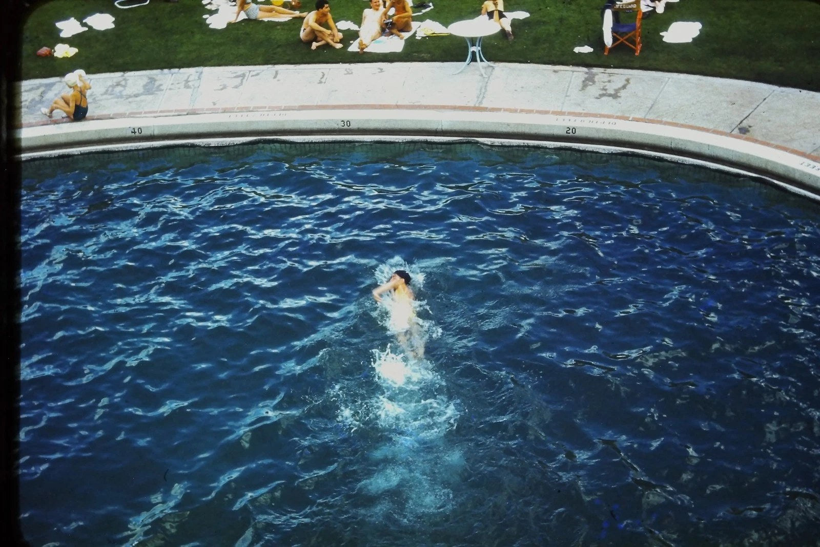 RED BORDER SLIDE Person splashing in blue pool, people lounging on grass.